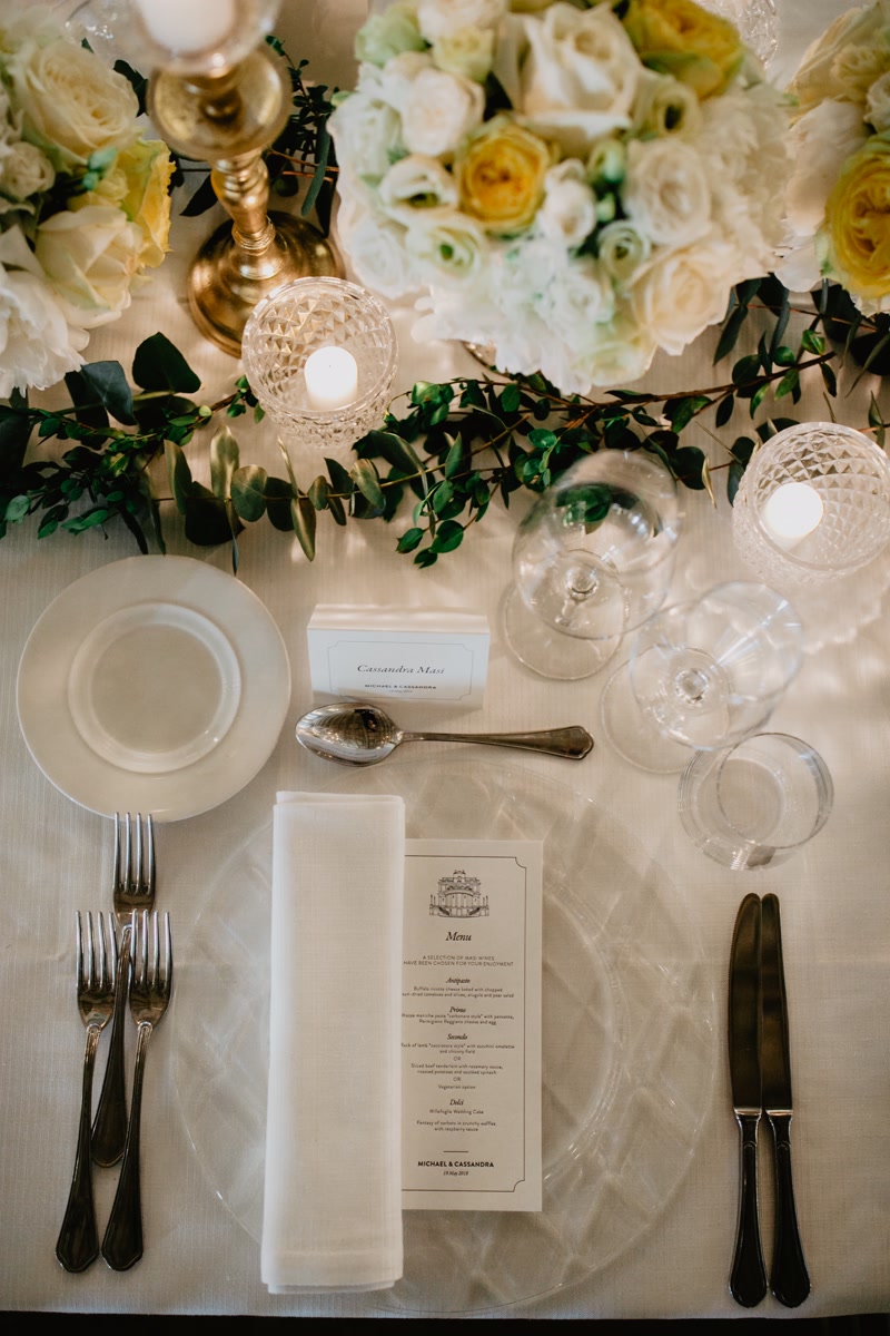 Printed menu, transparent glass chargers and white flowers as wedding table decor in Rome